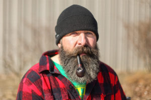 Rugged looking man in a toque and red plaid jacket, smoking a pipe