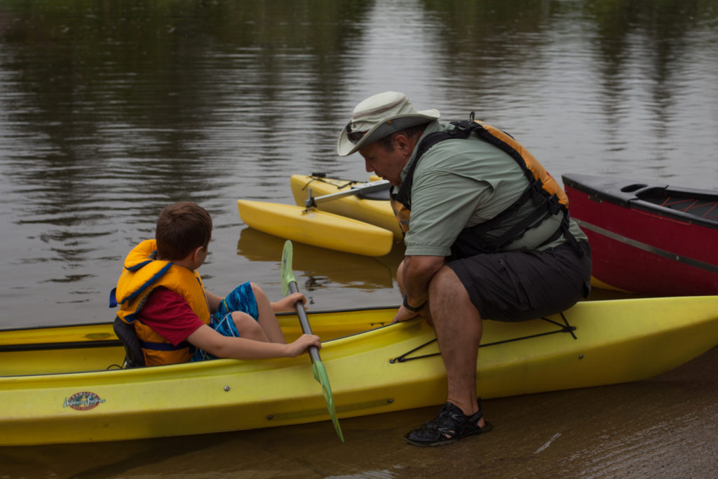 A mentor sits on a kayak, instructing a small boy to paddle